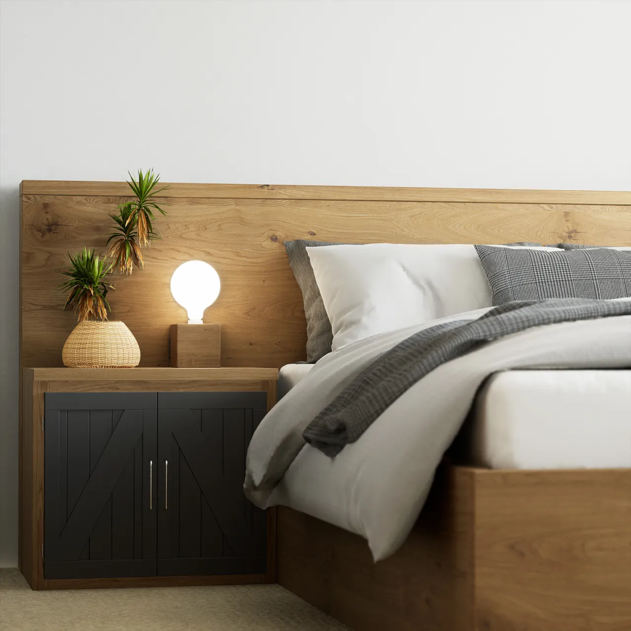 A bedroom corner in daylight. An oak headboard panel runs the width of the frame behind white and grey bedding with a checked throw; at the left a black slatted nightstand carries a rattan vase of dracaena and a bare globe bulb on an oak block, lit warm against a plain white wall.