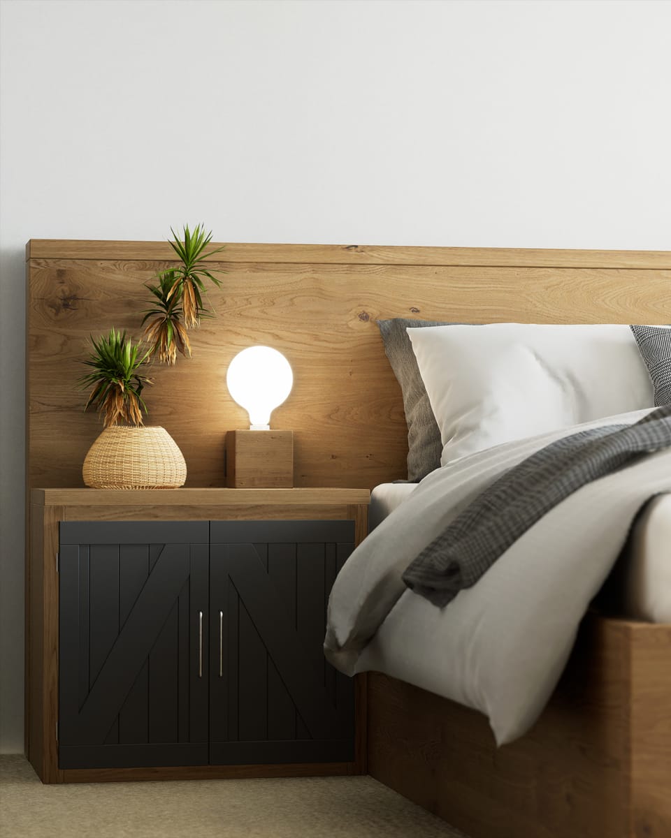 A bedroom corner in daylight. An oak headboard panel runs the width of the frame behind white and grey bedding with a checked throw; at the left a black slatted nightstand carries a rattan vase of dracaena and a bare globe bulb on an oak block, lit warm against a plain white wall.