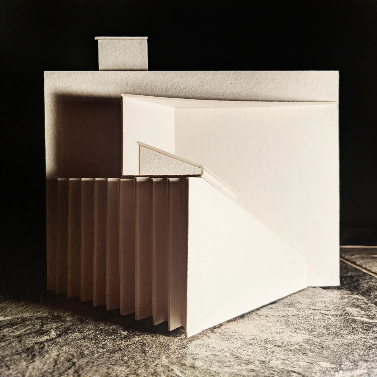 A photograph of a chipboard study model lit hard from the left against black. A flat roof plane overhangs a recessed void, a fine card handrail runs down a ramp, and a row of deep vertical fins throws a fan of shadows.