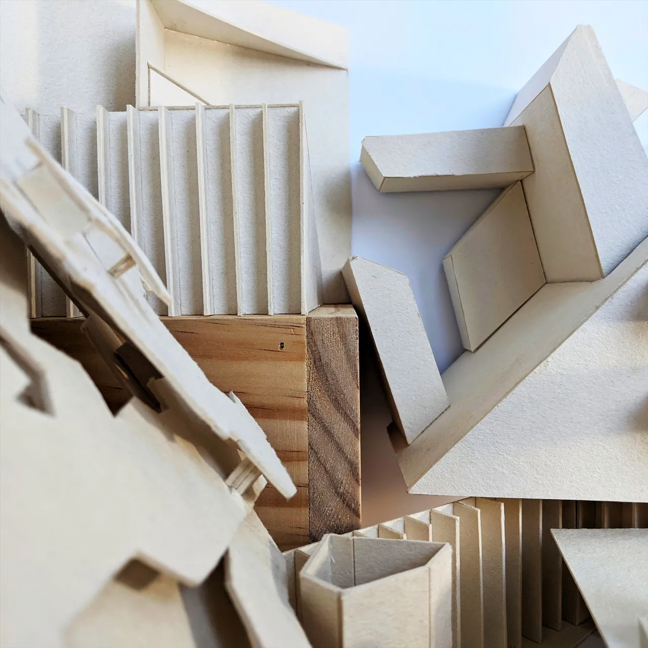 A close photograph into a cluster of chipboard model fragments (folded plates, finned walls and open trays) with a single block of raw timber wedged among the pale card.