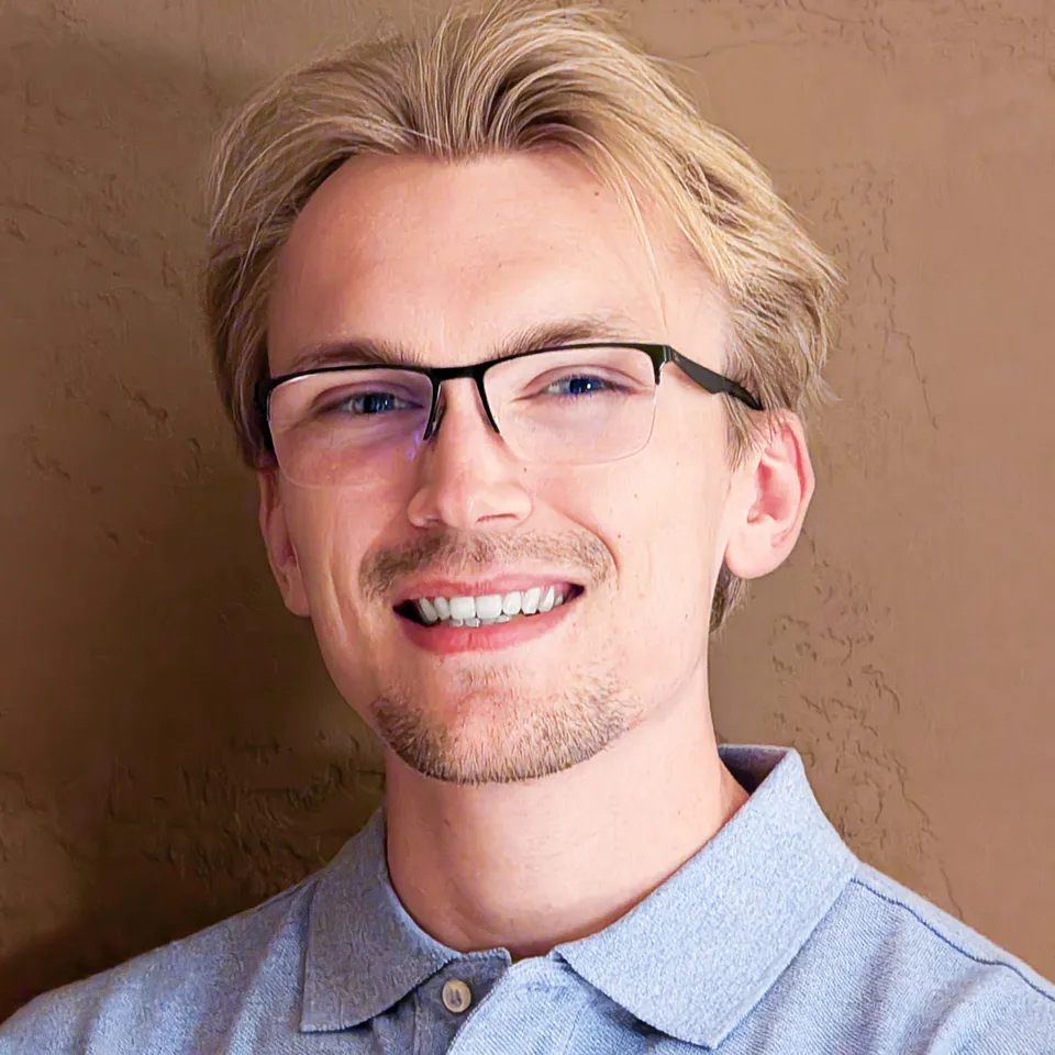 Coulter Heiberger, smiling, in glasses and a light blue collared shirt against a warm stucco wall.